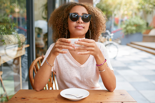 Portrait of pretty young woman sitting at sidewalk cafe drinking coffee. Attractive african woman having a cup of coffee.