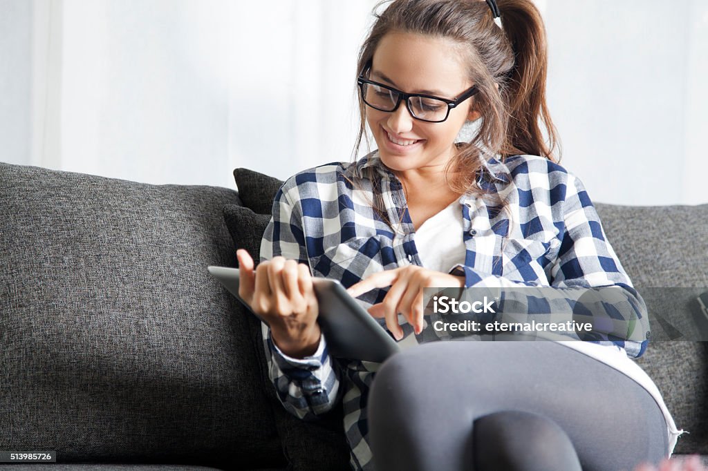 Using tablet Young woman sitting on sofa in living room and using tablet. Young Women Stock Photo