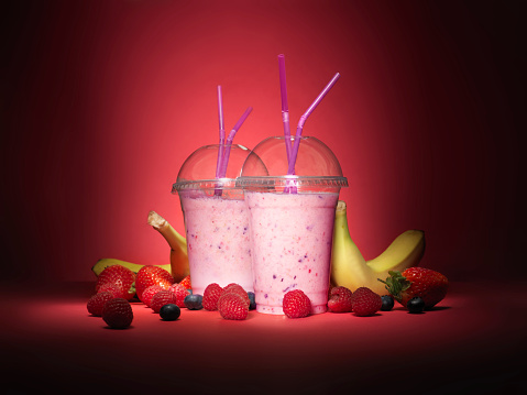 Shot of two take away smoothies in plastic cups on a red background with an array of different summer fruits spread around the shot.