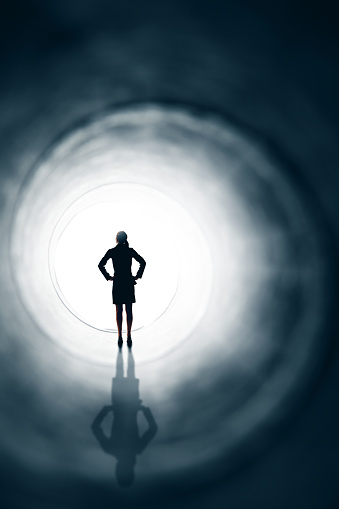 The silhouette of a business woman with her hands on her hips standing in front of a bright light at the end of a tunnel.