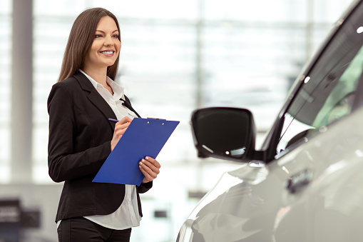 Beautiful young woman in classic suit is smiling and taking notes while examining car in a motor show
