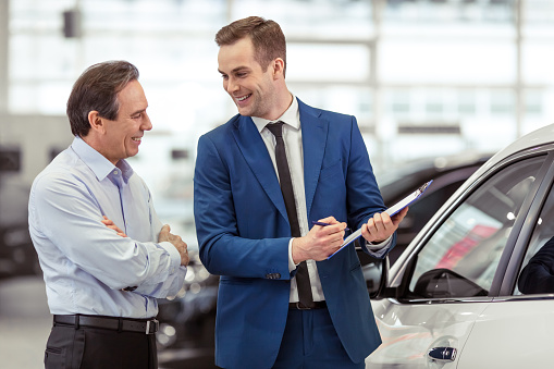 Handsome young man in classic blue suit is smiling while showing a car to middle aged customer in a motor show