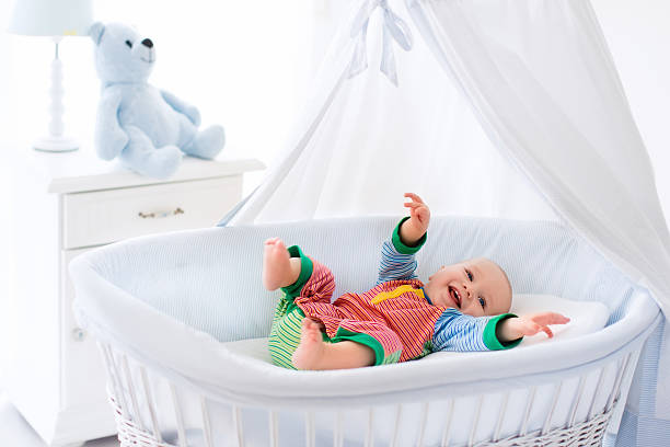 Funny baby in white crib with canopy. Nursery interior and bedding for kids. Laughing little boy playing in moses basket. Bedroom with bassinet for young children. Happy child in colorful pajamas.