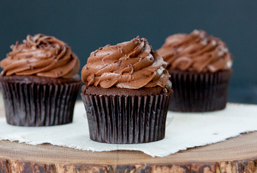 Three chocolate cupcakes with chocolate icing and chocolate sprinkles on cloak on a wood slice witha  black background