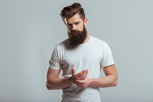 Handsome young bearded man is massaging his hand and looking at camera while standing against gray background
