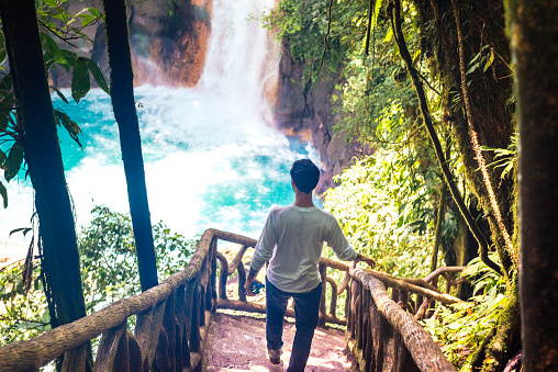 This is a horizontal, color, royalty free stock photograph of a 39 year old Cuban man walking a wooden staircase as he descends toward the eco tourist, travel destination, Rio Celeste, a waterfall in Tenorio National Park, Costa Rica. Photographed with a Nikon D800 DSLR camera.