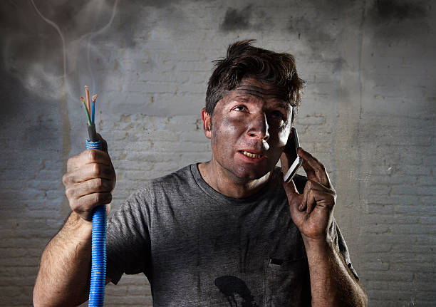 electrocuted man calling for help in dirty burnt funny face young man holding electrical cable smoking after electrical accident with dirty burnt face in funny desperate expression calling with mobile phone asking for help in electricity DIY repairs danger concept terrified photos stock pictures, royalty-free photos & images