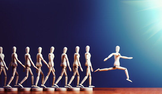 A single wooden lay figure leaps ahead of a line of other marionettes into the spotlight. Copy space on the dark blue background.