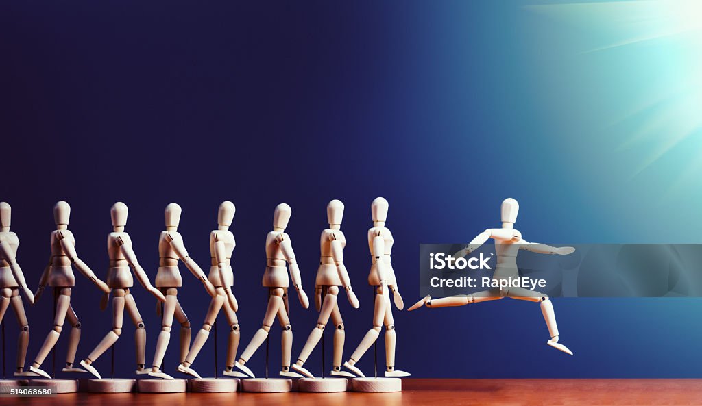 Brave puppet breaks away from pack, leaping into spotlight A single wooden lay figure leaps ahead of a line of other marionettes into the spotlight. Copy space on the dark blue background. Initiative Stock Photo