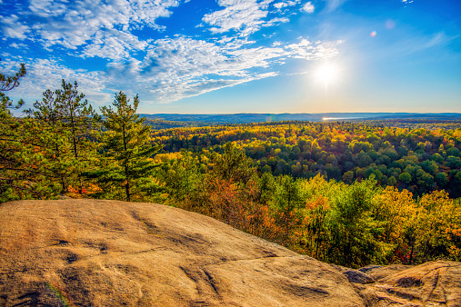 A high angle view of the colorful autumn treetops viewed from above on a rocky cliff.  Golden sunlight shines down on the landscape with soft clouds in a blue sky.
