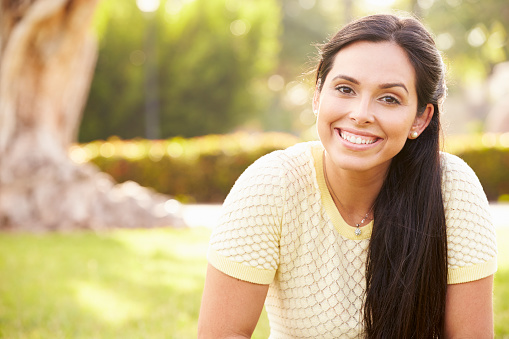 Portrait Of Young Hispanic Woman Sitting In Park Smiling To Camera