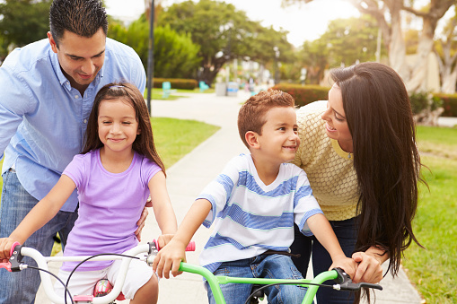 Parents Teaching Children To Ride Bikes In Park Smiling