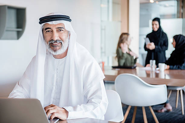 Mature Arab businessman wearing ghutra using laptop, portrait Businessman working in office with laptop computer wearing traditional Arabian agal and ghoutra. middle eastern culture photos stock pictures, royalty-free photos & images