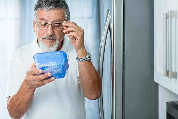 Senior man in his kitchen by the fridge Is this still fine? Senior man in his kitchen by the fridge, looking at the expiry date of a product she took from her fridge - obsolete stock pictures, royalty-free photos & images