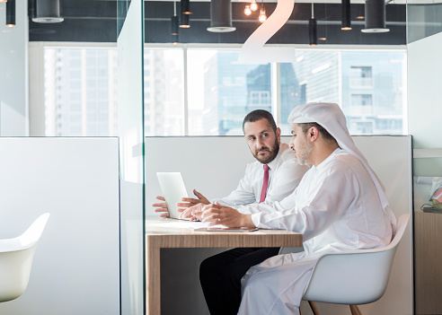 Middle Eastern businessmen in business meeting in open plan workspace in modern office one is in traditional Arab kandura while the other is wearing shirt and tie.