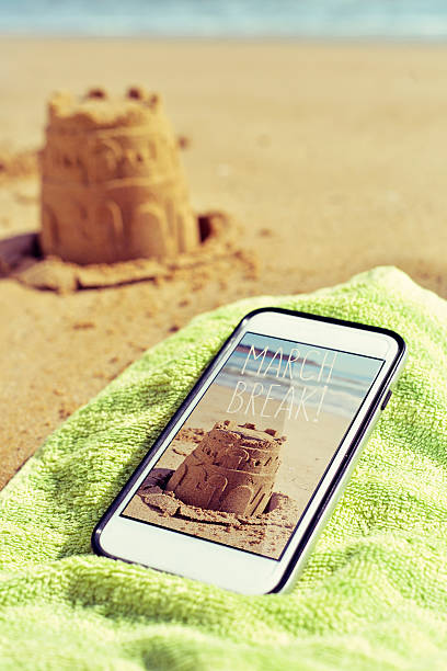 a smartphone with a picture shot by myself of a sandcastle and the text march break in its screen, placed on a green towel on the sand of a beach and the depicted sandcastle in the background