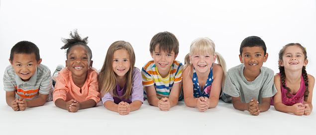 A multi-ethnic group of elementary age students are lying in a row and are smiling while looking at the camera. The photo is isolated on white.