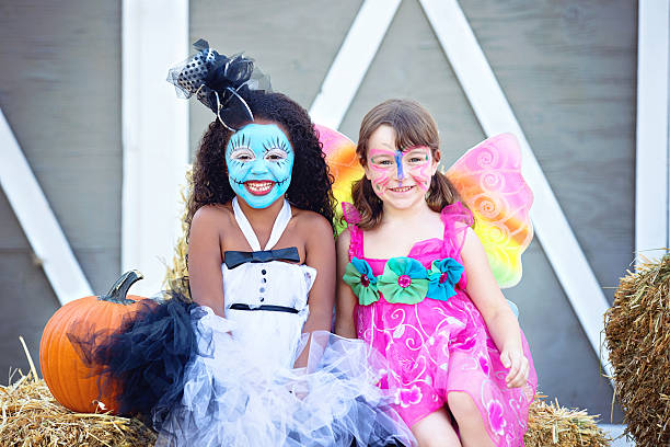 Two Girls In Costume at A Halloween Party Two adorable girls dressed up as a doll and a butterfly sitting on a bale of straw at a Halloween party. halloween face paint stock pictures, royalty-free photos & images