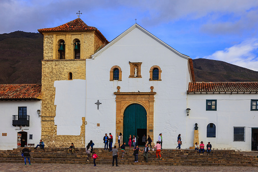 Villa de Leyva, Colombia - September 13, 2014: Founded in 1572 and located at just over 7000 feet above sea level on the Andes Mountains, Villa de Leyva was declared a National Monument in 1954 to protect it's colonial architecture and heritage. It is located in the Department of Boyaca, in the South American country of Colombia.  Photo shows the church on the main plaza or square.  People visiting the town and local residents are seen on the square just sitting and relaxing, chatting with friends or going about their routine chores. Photo shot in the late afternoon sunlight; horizontal format. Copy space.