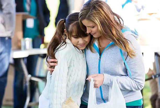 Woman embraces daughter as they receive donations at outdoor community clothing drive. The woman is holding a plastic bag containing donated items.