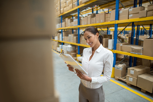 Asian business woman working at a warehouse doing an inventory and holding a clipboard
