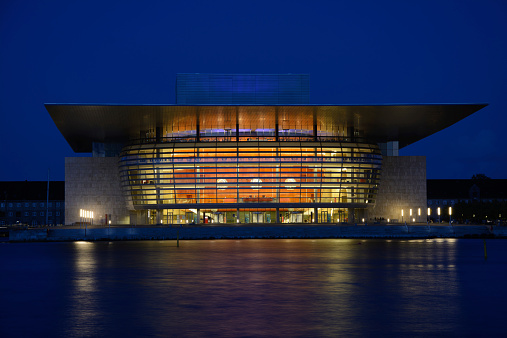 The Opera (Operaen) building in Copenhagen photographed in the blue hour of dusk. The combination of the spectacular lamps in the foyer, the wooden covering of interior and the reflections in the water makes a grand sight. The building was photographed slightly of center to make it really stand out.