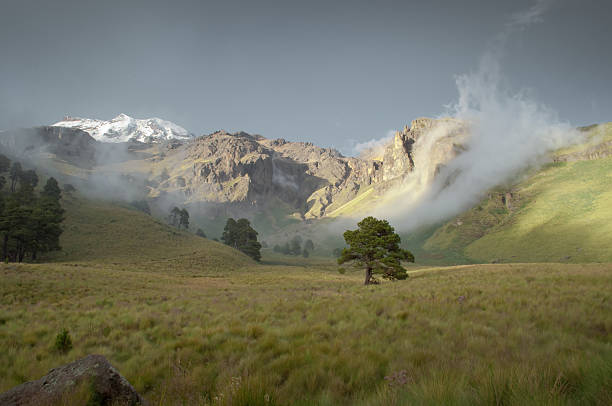 Popocatepetl volcano eruption smoke at the daytime among other green hills and mountains, with cloudy blue sky