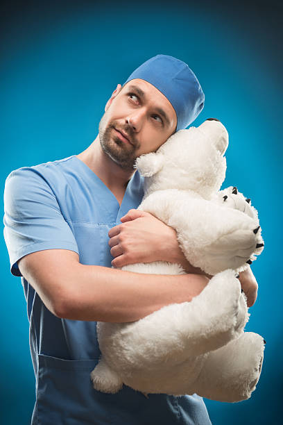 Hard work of doctors Half-length portrait of dark-haired lovely doctor wearing blue medical uniform keeping a big white teddy bear thinking about his girlfriend. Isolated on blue background man with teddy bear stock pictures, royalty-free photos & images