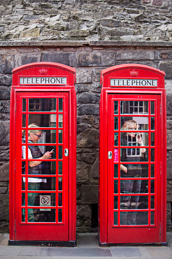 Two women inside a telephone booth talking and text with the mobile phones.