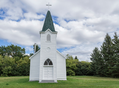 horizontal image of a quaint little white country church with a steeple sitting in a green meadow surrounded by trees under a beautiful blue sky with white clouds floating by in the summer time.