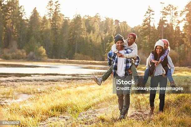 Parents Giving Children Piggyback Ride On Walk By Lake Stock Photo - Download Image Now