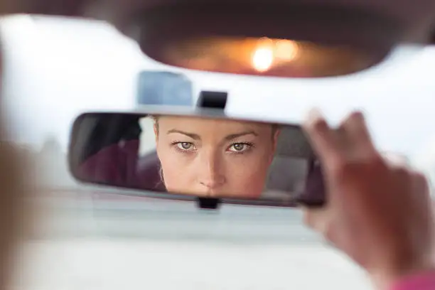 Beautiful young lady looking back through the rear view mirror from the front seat of a car while reversing.