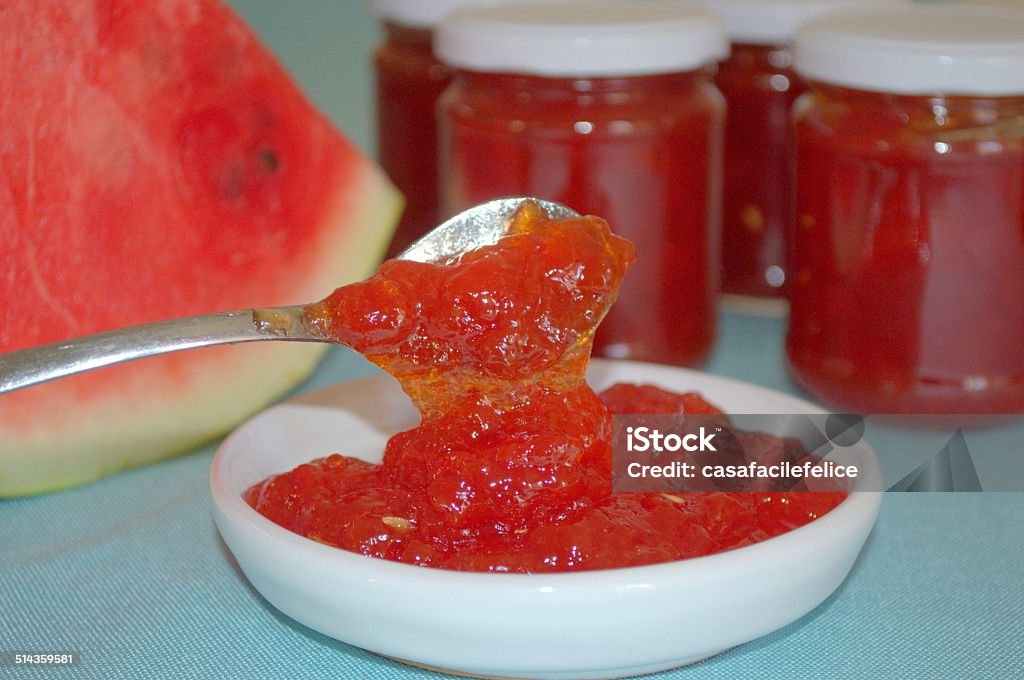Watermelon Jam On the right two jars full of watermelon jam; on the left slice of watermelon; in the foreground watermelon jam on a white plate and other that slides from a spoon. Watermelon Stock Photo