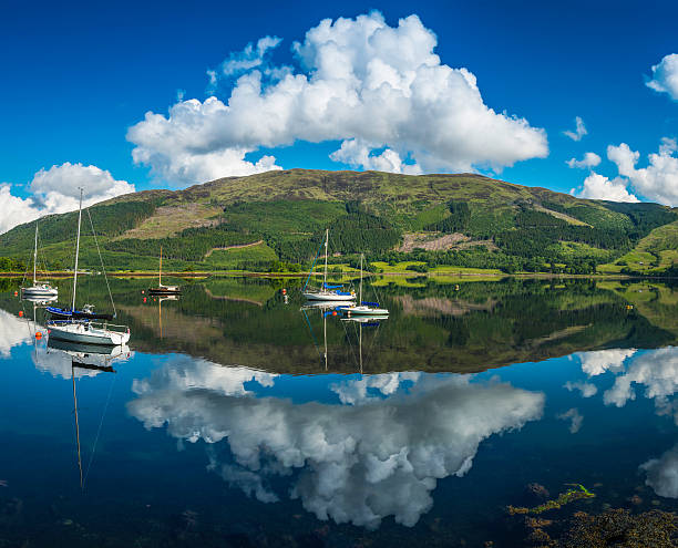 White fluffy clouds, green hills, blue sky and moored yachts reflecting in the perfectly still waters of Loch Leven at Glen Coe deep in the Highlands of Scotland, UK. ProPhoto RGB profile for maximum color fidelity and gamut.