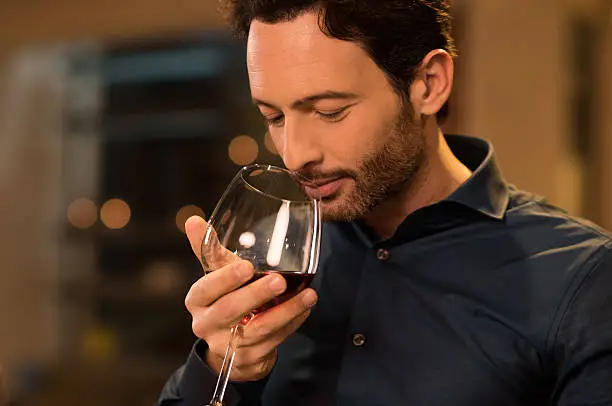 Handsome young man smelling red wine before drinking it. Young man drinking red wine in a luxury restaurant. Handsome young man tasting a glass of red wine during dinner.