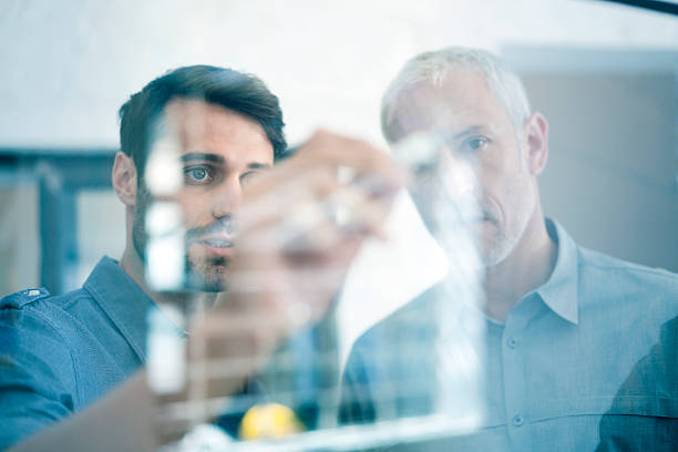 Businessmen making plan on glass wall in office A photo of businessmen making plan on glass wall. Concentrated executive is explaining strategy to colleague. Professionals are at workplace. business plan stock pictures, royalty-free photos & images
