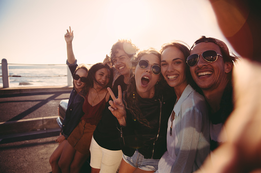 Group of hipster friends cheering and showing peace sign while taking a self portrait close to the beach on a summer afternoon