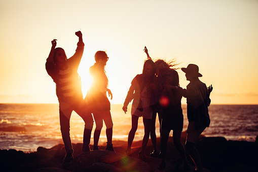 Group of fun loving hipser friends dancing with raised arms and partying at sunset on a beach in summertime