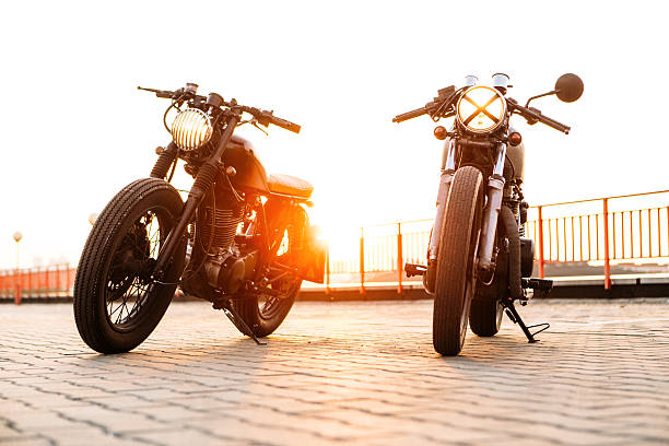 Two black and silver vintage custom motorcycles caferacers Front view of two vintage custom motorbike caferacer motorcycle one with grill headlight another with tape cross on empty rooftop parking lot with backlight sun during sunset. Hipster lifestyle. cafe racer stock pictures, royalty-free photos & images