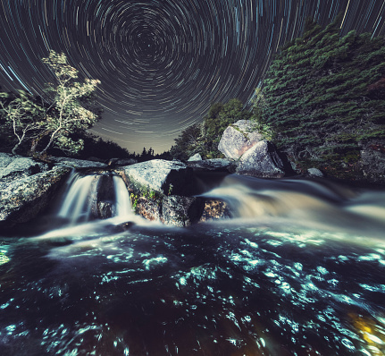 A river cascades over a granite landscape under a canopy of spinning stars.  Long exposure with light painting.