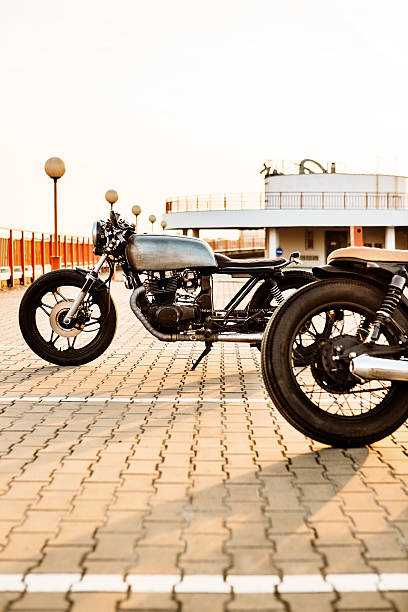 Two vintage custom motorcycle caferacer motorbikes on empty rooftop parking lot surrounded by urban environments midtown buildings during sunset. Hipster lifestyle, student dream.