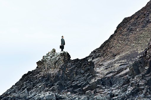 A businessman, holding his briefcase, stands on a rocky slope and looks up at the challenge of an even steeper slope in front of him.