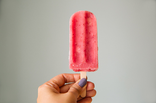 Woman holds up a pink strawberry popsicle in front of a blue background