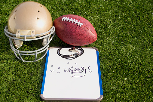 A gold helmet, football and clipboard on a field.