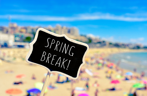 closeup of a black signboard with the text spring break written in it, in front of a blurred beach with many people bathing and sunbathing