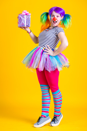 Cheerful young woman in a multi-colored wig, dressed in a clown costume. She is holding in her hand a brightly wrapped gift. Studio shooting on a yellow background