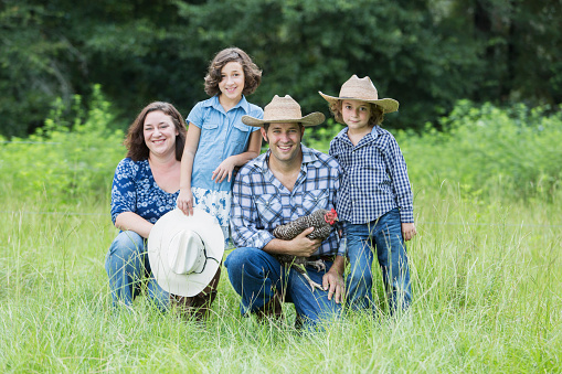 A family with two children on a farm, posing in a field, smiling at the camera.  The father is holding a chicken in his arms. They are wearing jeans, cowboy hats and plaid shirts.
