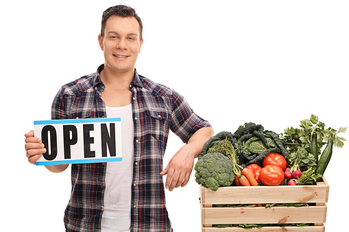 Young market vendor holding an open sign next to a crate full of vegetables isolated on white background