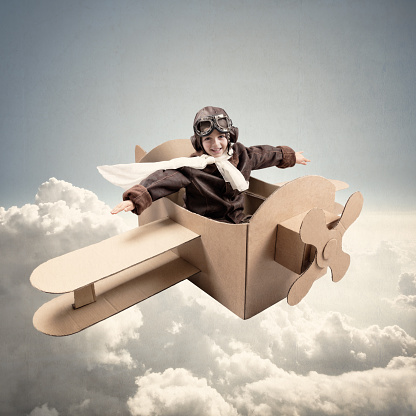 Boy wearing old-fashioned aviator hat, scarf and goggles flying a cardboard airplane in his imagination