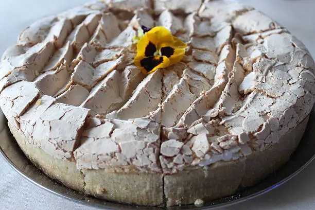 Photo showing a homemade blueberry cooked cheesecake with a crunchy meringue topping, decorated with a yellow edible pansy flower and cut into slices, ready to be served.